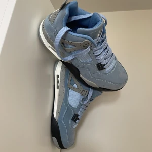 Nike Air Jordan 4 University Blue - Nike Air Jordan 4 University Blue med ljusblå mocka och mesh, grå detaljer med svarta stänk och svart/vit sula. Ikonisk Jumpman-logga på plösen och klassisk snörning. Perfekta för dig som vill sticka ut med stilrena sneakers.