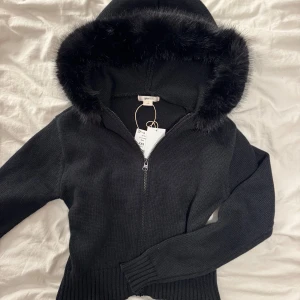 Svart fur hoodie från Gina Tricot - Säljer en helt ny svart hoodie från Gina Tricot med fluffig päls runt huvan. Tröjan är stickad, har dragkedja framtill och ribbade muddar vid ärmslut och nederkant. Perfekt för dig som vill ha en mysig och snygg look under hösten och vintern.💖