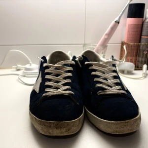 Golden Goose Superstar sneakers blå - Golden Goose Superstar sneakers i mörkblå mocka med vit stjärndetalj på sidan och vit häl. Skorna har vita snören och en platt sula med vintage-look. Insidan är vit och har tydlig branding. Perfekt för dig som gillar coola och unika sneakers med streetkänsla. Lite smutsiga säg till om jag ska borsta till mockan så fixar jag det! Box och dustbag finns👍Kan passa 44 bara några månader gamla så sulan in bra skick