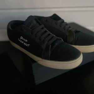 Svarta Saint Laurent sneakers - Svarta sneakers från Saint Laurent med vit logga på sidan och vit sula. Skorna har snörning och en klassisk låg profil. Materialet är en mix av skinn och textil, vilket ger en stilren och modern look. Perfekta för dig som gillar exklusiva och trendiga sneakers.