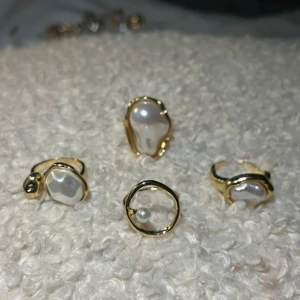 Guldiga ringar med pärldetaljer - Fyra unika ringar i guldton med oregelbundna former och stora vita pärlor. En av ringarna har en rund ram med en liten pärla i mitten, medan de andra har organiska, vågiga kanter som omsluter pärlorna. Perfekt för dig som gillar statement-smycken med lyxig vibe.