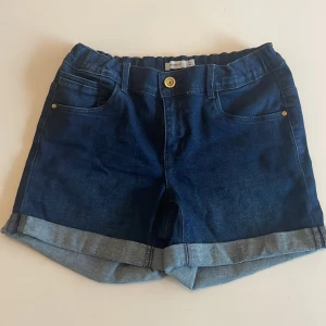 Blå jeansshorts från Name It💕💫 - Snygga mörkblå jeansshorts från Name It i storlek 164. Modellen har uppvikta benslut med dekorativa detaljer i guld, klassiska fickor fram och bak samt gylf med knapp. Perfekta för sommaren och har en normal passform. Använda max fem gånger pga de inte riktigt passade min stil. 💋✨🤩