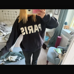 Svart sweatshirt med PARIS-tryck - Cool svart sweatshirt med stort vitt PARIS-tryck på bröstet. Tröjan har en loose passform och långa ärmar, perfekt för chill dagar. Mjuk bomullskänsla och klassisk rund hals. Enkel att matcha med jeans eller leggings för en avslappnad vibe.