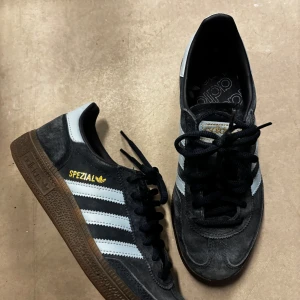Adidas Spezial sneakers i mörkblå mocka - Säljer ett par Adidas Spezial sneakers i mörkblå mocka med klassiska blåa ränder på sidorna och gummisula. Skorna har svart snörning, guldfärgad Spezial-logga och rund tå. Perfekta för dig som gillar retrostil och streetwear.