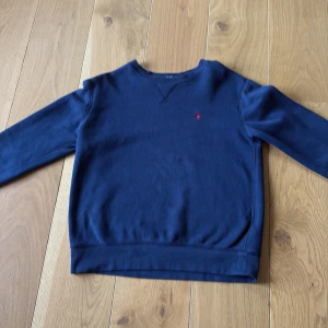Mörkblå tröja från Ralph Lauren - Klassisk mörkblå sweatshirt från Ralph Lauren med liten röd logga broderad på bröstet. Tröjan har ett pyttelitet hål på framsidan som inte syns och två små fläckar på ärmen som bara syns på nära håll, därför är tröjan så billig