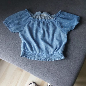 Blå blommig blus med puffärm - Supersöt ljusblå blus från Kappahl med små vita blommor, puffiga korta ärmar och smockad midja. Blusen har offshoulder-modell och volangdetaljer vid halsringning och ärmslut. Perfekt för vår och sommar!