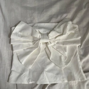 Vit topp med stor rosett från Kappahl - Supersöt vit topp från Kappahl med en stor rosett framtill som verkligen sticker ut. Toppen har dragkedja i sidan och är gjord i ett slätt tyg som känns fräscht och stilrent. Perfekt för dig som vill ha något unikt och trendigt i garderoben.