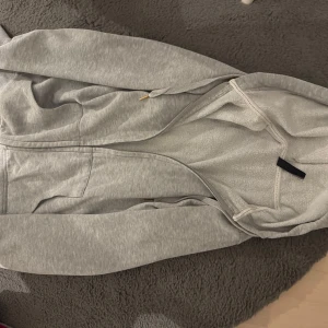 Grå hoodie från Colors of the World - Säljer en ljusgrå hoodie från Colors of the World i storlek S. Tröjan har huva med snörning, dragkedja framtill och två fickor. Mjuk insida och klassisk passform, perfekt för chill dagar. Enkelt och stilrent plagg som funkar till det mesta.