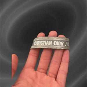 Grått armband från Christian Dior - Snyggt grått armband från Christian Dior med vit broderad logga. Armbandet har en enkel och stilren design som passar perfekt till din vardagsstil. Tillverkat i textilmaterial med tydlig branding.
