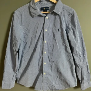 Randig skjorta från Ralph Lauren - Klassisk skjorta från Ralph Lauren i blåvitt randigt mönster. Skjortan har lång ärm, knappar framtill och på manschetterna samt den ikoniska broderade loggan på bröstet. Tillverkad i mjuk bomull och har en normal passform. Det är Xl i barn storlek men skulle säga S