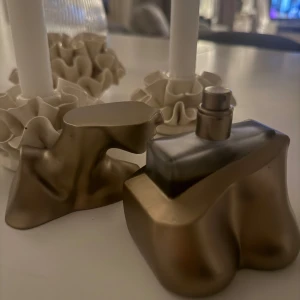 Parfym  - Unik parfymflaska i form av en stiliserad kvinnlig byst i matt guld. Flaskan har en konstnärlig och modern design med mjuka former och är tillverkad i glas och metall. Perfekt som statement-piece på sminkbordet.
