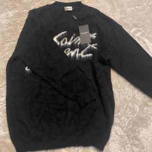 Svart Saint Laurent stickad tröja - Säljer en svart stickad tröja från Saint Laurent med vit, fluffig logga framtill. Tröjan har lång ärm och en klassisk rund halsringning. Perfekt för dig som vill ha en statement piece med lyxig känsla och cool design.