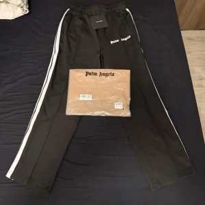 Palm Angels svarta trackpants - Svarta trackpants från Palm Angels med vita sidoränder och logga tryckt på vänster höft. Byxorna har elastisk midja och raka ben, perfekt för en streetwear-look. Snygga detaljer och klassisk design som sticker ut. Passar storlek M!!!!