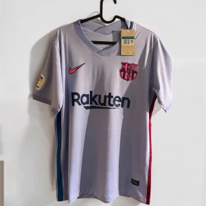 FC Barcelona Nike fotbollströja lila - FC Barcelona matchtröja från Nike i ljuslila med klubbmärke och Rakuten-tryck på bröstet. Kortärmad modell med La Liga-märke på ärmen och Unicef-tryck på ryggen. Tillverkad i lätt och ventilerande material, perfekt för fotbollsplanen.