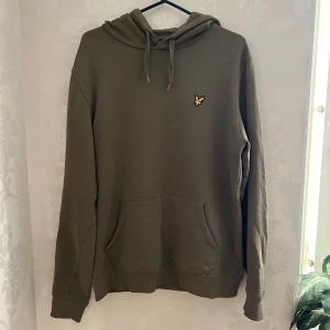Olivgrön hoodie från Lyle & Scott - Säljer en olivgrön hoodie från Lyle & Scott med klassisk gul logga på bröstet. Tröjan har huva med snörning, magficka och långa ärmar. Perfekt för dig som gillar en enkel och stilren look.