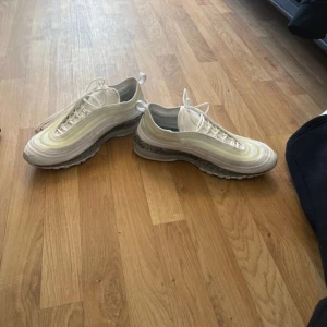Nike Air Max 97 vita sneakers - Nike Air Max 97 sneakers i vitt med ljusgula detaljer och reflekterande inslag. Klassisk vågig design, synlig Air Max-sula med stänk i grått och transparent. Ovandel i syntet och mesh, snörning och dragflik bak. Perfekta för dig som gillar streetstyle.