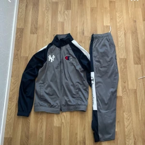 Grå och marinblå Champion tracksuit - Snygg tracksuit från Champion i grått och marinblått med vita detaljer. Jackan har dragkedja, New York Yankees-logga och Champion-märke på bröstet. Byxorna har elastisk midja och Champion-logga på benet. Perfekt för chill eller sport.