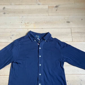 Marinblå skjorta från Polo Ralph Lauren - Snygg marinblå skjorta från Polo Ralph Lauren med klassisk krage och knäppning framtill. Skjortan har långa ärmar och den ikoniska broderade loggan på. Perfekt för en stilren och avslappnad look.