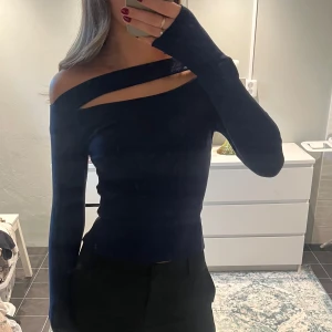 Mörkblå offshoulder ribbad topp - Säljer en mörkblå, ribbad topp med lång ärm och offshoulder-design. Toppen har en cool symmetrisk cutout vid halsen som ger en unik look. Sitter tight och framhäver figuren, perfekt för dig som gillar trendiga detaljer.