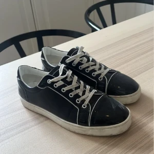 Svarta sneakers med röd sula - Snygga svarta sneakers, i glansigt skinn med vita kontrastsömmar och snörning. Ikonisk röd sula och Louboutin-signatur på hälen. Perfekta för dig som vill sticka ut med exklusiva detaljer och streetstyle-vibe. Lite uppslitet vid sidan av skon och lite slitet på sulan som ni ser på bilden