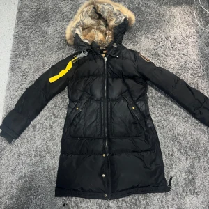 Svart lång dunjacka Parajumpers XS - Säljer en svart lång dunjacka från Parajumpers med fuskpälsfodrad huva och ikonisk gul rem på axeln. Jackan har dragkedja, knappar och flera fickor framtill. Perfekt för kalla vinterdagar och har snygga detaljer som logopatch på ärmen.