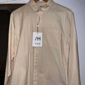 Beige skjorta från Selected Homme - Snygg beige skjorta från Selected Homme i klassisk modell med button-down krage och bröstficka. Skjortan är tillverkad i mjuk bomull och har långa ärmar med knappar vid manschetterna. Perfekt för dig som gillar stilrena och enkla plagg. Aldrig använd och prislapp sitter kvar. 