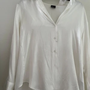 Vit satinliknande skjorta Gina Tricot - Snygg vit skjorta från Gina Tricot i satinliknande material med glansig finish. Skjortan har v-ringad krage, långa ärmar och knappar framtill. Passformen är loose och den har en något längre och rundad nederkant. Perfekt för dig som gillar stilrena och trendiga plagg.