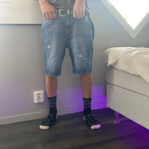 Blå jeansshorts från 157 - Säljer ett par klassiska blå jeansshorts från 157 med normal passform och midwaist. Shortsen har fem fickor, bälteshällor och slitna detaljer framtill för en chill vibe. Perfekta för sommardagar och hänger snyggt strax ovanför knäna.