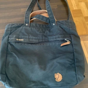 Marinblå shoppingväska Fjällräven G-1000 - Säljer en marinblå shoppingväska från Fjällräven i slitstarkt G-1000-material. Väskan har både handtag och axelremmar, en dragkedjeficka framtill och klassisk läderdetalj med logga. Perfekt för dig som vill ha en rymlig och stilren väska till vardagen. Kan även användas som ryggsäck 