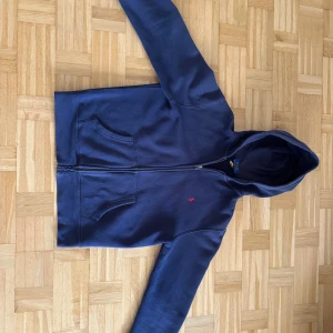Mörkblå hoodie från Polo Ralph Lauren - Snygg mörkblå hoodie från Polo Ralph Lauren med dragkedja och huva. Tröjan har två stora fickor framtill och den klassiska röda Polo-loggan broderad på bröstet.