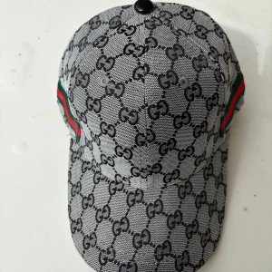 Gucci keps med GG-mönster - Snygg keps från Gucci med klassiskt GG-mönster i svart och grått. Kepsen har gröna och röda ränder på sidorna och justerbar rem i svart skinn. Tillverkad i 90% merinoull och 10% kashmir för lyxig känsla. Perfekt för dig som vill sticka ut med designerstil.