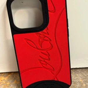 Röd och svart Christian Louboutin mobilskal - Snygg mobilväska från Christian Louboutin i rött och svart med ikonisk logga i relief. Utsidan är röd med svart kant och detaljer, baksidan är glittrigt svart. Perfekt accessoar för dig som vill sticka ut med lyxig vibe.