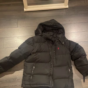 Svart pufferjacka från Polo Ralph Lauren - Säljer en svart pufferjacka från Polo Ralph Lauren med huva och dragkedjor på fickorna. Jackan har en klassisk röd logga på bröstet och är perfekt för kalla dagar. Stora quiltade partier och justerbara ärmslut ger extra komfort och stil. Kan tänkas sänka priset. Om man köper så får man med en svart polo mössa med röd polo logga 