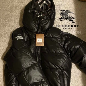 Burberry Jacka - Svart pufferjacka från Burberry med stor huva och klassiskt rutigt foder. Jackan har dragkedja, fickor och tydlig Burberry-logga på ärmen. Perfekt för kalla dagar när du vill ha både stil och värme.