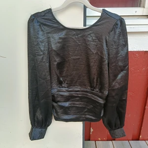 Svart blus med v-ringning och smock - Snygg svart blus i glansigt material med lång ärm och bred smockad midja. Djup v-ringning i ryggen och knappar vid ärmslut. Perfekt för dig som gillar stilrena och trendiga plagg med en twist.