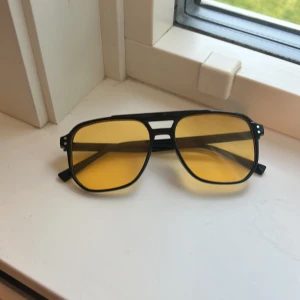 Svarta pilotglasögon med gula linser - Snygga pilotglasögon med svart ram och gula linser som ger en cool retrovibe. Ramen är i plast och har en klassisk dubbelsidig brygga. Perfekt för dig som vill sticka ut med en unik stil och få extra färg på din look.