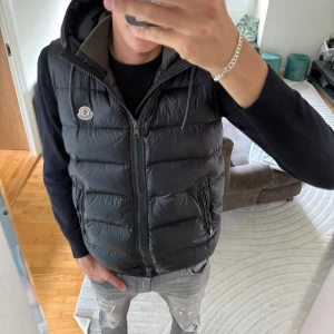 Svart dunväst från Moncler med huva - Säljer en svart dunväst från Moncler med huva och dragkedja framtill. Västen har två fickor med dragkedja och klassisk Moncler-logga på bröstet. Den är quiltad och har en stilren, sportig look som passar perfekt till lager-på-lager-stil.