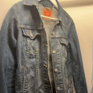 Levi's jeansjacka blå - Säljer en klassisk blå jeansjacka från Levi's med snygga sömmar, bröstfickor med knappar och silverfärgade knappar framtill. Jackan har en tidlös design med krage och långa ärmar. Perfekt för dig som vill ha en ikonisk denimlook. 💙