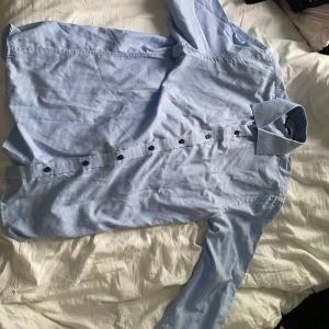 Blårutig skjorta från Dressmann L - Snygg blårutig skjorta från Dressmann i storlek L. Skjortan har klassisk krage, långa ärmar och mörka knappar som kontrast. Perfekt för dig som gillar stilrena och tidlösa plagg. Tillverkad i mjuk bomull för en skön känsla hela dagen.