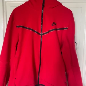 Röd Nike-tech Zip Hoodie - Nike zip hoodie i stark röd färg med svart logga på bröstet och svarta detaljer längs bröstet. Hoodien har hel dragkedja, huva och en praktisk ficka med dragkedja på ärmen. Perfekt för dig som gillar sportig och clean stil.