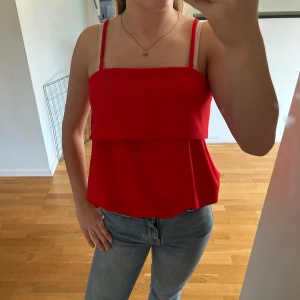 Röd linnetopp med volang - Snygg röd linnetopp med  smala axelband och volangdetalj över bysten. Toppen har rak halsringning och ärmlös design, perfekt till jeans eller kjol för en trendig look.