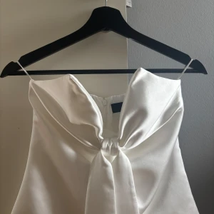 Vit axelbandslös topp med rosett - Säljer en elegant vit axelbandslös topp i satinliknande material. Toppen har en stor rosett framtill. Perfekt för dig som vill sticka ut med en stilren och chic look. Dragkedja baktill för enkel påklädning.