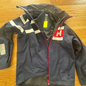Helly Hansen marinblå seglar jacka M - Säljer en marinblå vindjacka/seglar jacka  från Helly Hansen i storlek M. Jackan har vita detaljer, röd dragkedja och stor HH-logga på bröstet. Perfekt för blåsiga dagar och har en sportig look med praktiska fickor och huva. Pris är diskuterbart 