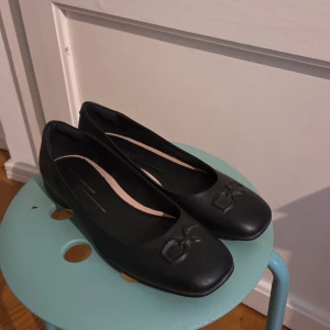 Svarta ballerinaskor från ECCO - Stilrena svarta ballerinaskor från ECCO i mjukt skinn. Skorna har en fyrkantig tå och en snygg präglad detalj framtill. Perfekta för dig som gillar klassisk och enkel design med en twist. Lätta att matcha till olika outfits.