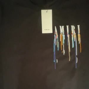 Svart Amiri t-shirt med färgstänk - Svart t-shirt från Amiri i storlek L med stort tryck framtill där AMIRI-loggan har färgstänk i blått, orange, vitt och rött som rinner ner. Snygg och modern design med cool streetkänsla. Rund hals och klassisk passform.