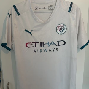 Manchester City vit matchtröja Puma M - Säljer en officiell Manchester City fotbollströja från Puma i vitt med mörkblå detaljer. Tröjan har klubbmärke på bröstet, sponsortryck från Etihad Airways och Nexen Tire samt klassisk V-ringning. Tillverkad i lätt och ventilerande funktionsmaterial.