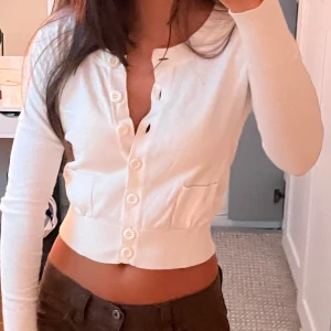 Vit croppad kofta från New Look - Supersöt vit croppad kofta från New Look med långa ärmar och stora knappar framtill. Tröjan har två små fickor och är i mjukt, stretchigt material som sitter tajt och snyggt. Perfekt till jeans eller kjol för en clean och fräsch look.