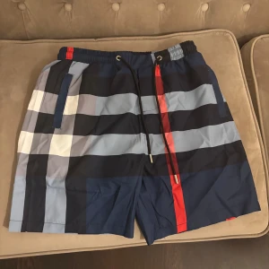 Burberry shorts - Helt nya och oanvända shorts från Burberry med breda ränder i marinblått, ljusblått, grått, vitt och en röd detalj. De har elastisk midja med snörning och två sidofickor. Perfekta för sommaren och har en modern, loose passform.
