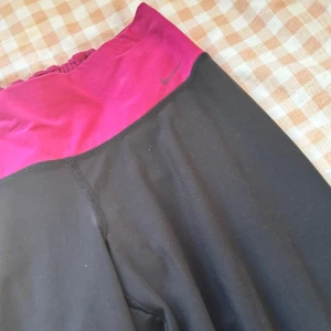 Nike svarta träningsbyxor med lila midja - Svarta träningsbyxor från Nike med bred lila midja och diskret logga framtill. Byxorna har bootcut-modell och är tillverkade i ett stretchigt syntetmaterial som passar perfekt för träning eller chill. Snygg passform och bekväm känsla.