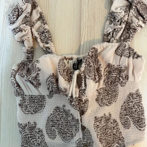 Beige smockad topp med paisleymönster H&M - Supersöt smockad topp från H&M i beige med brunt paisleymönster. Toppen har breda, rynkade axelband, knappar framtill och en liten knytning vid bysten. Perfekt för dig som gillar boho-vibbar och vill ha något unikt i garderoben. Helt oanvänd. Nypris: 229:-